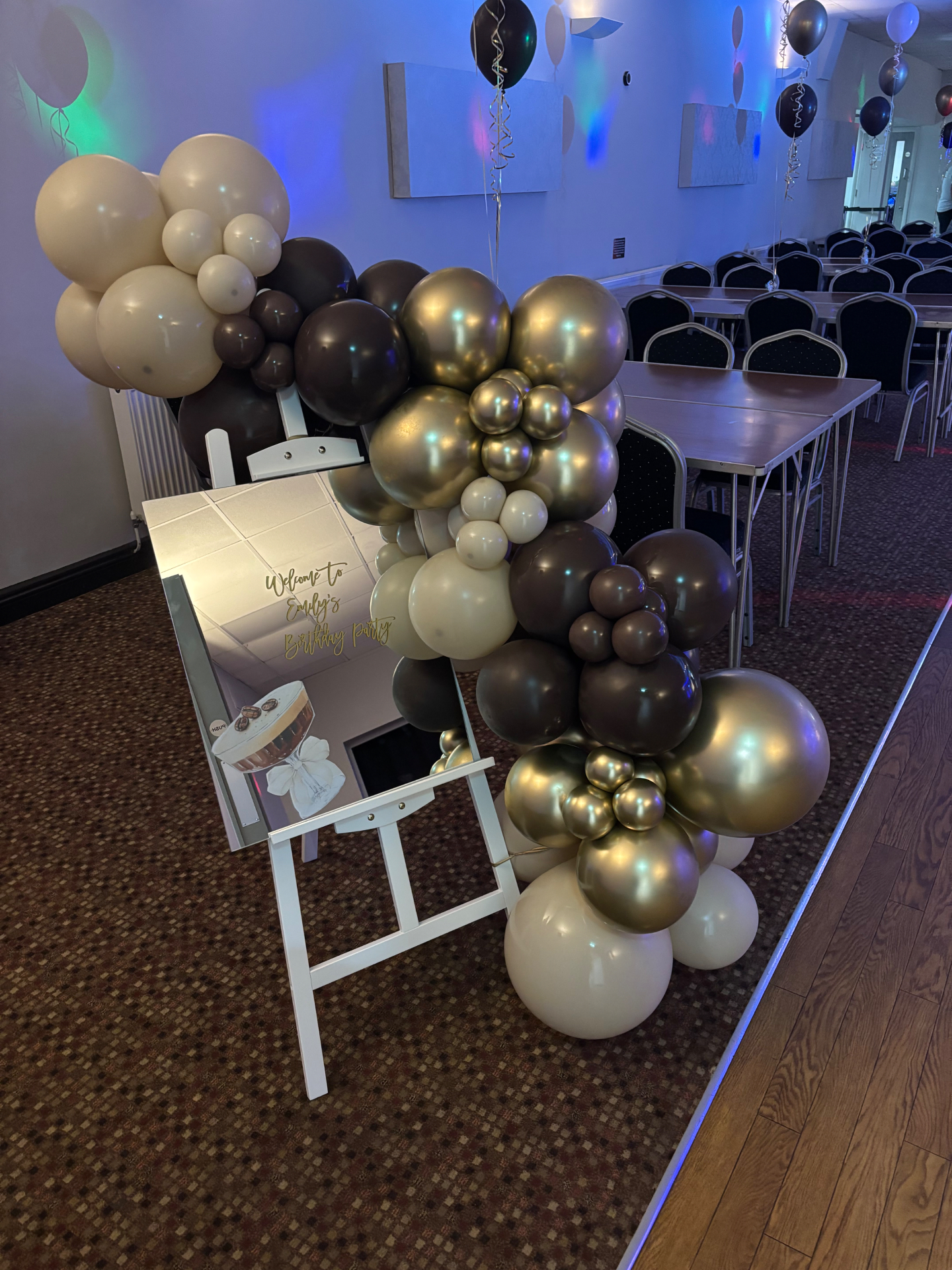 Personalised mirrored welcome sign on a white easel with a cream, gold and brown balloon garland at a birthday party.