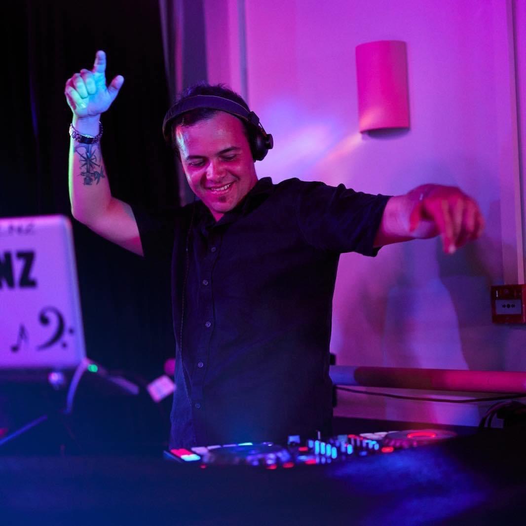 DJ T.L.K performing live with vibrant magenta and blue lighting at an event, wearing headphones behind the decks.